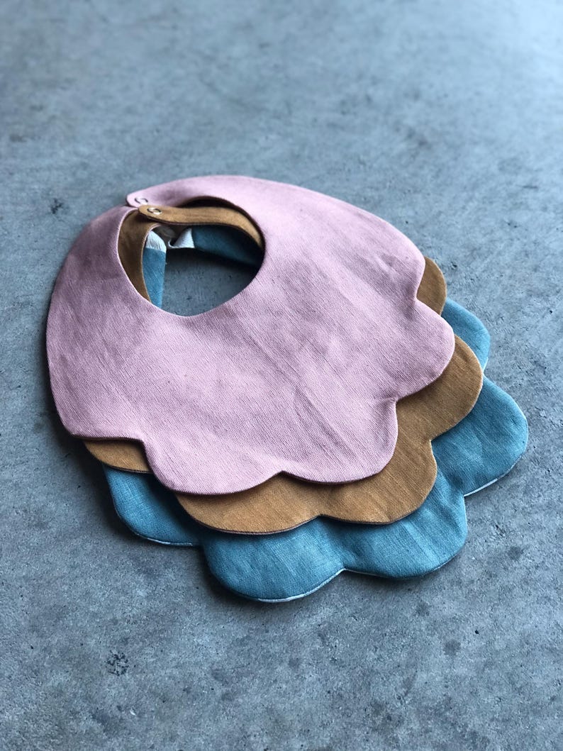 May include: Three scalloped-edge baby bibs in pink, brown, and blue. The bibs are made of a soft, textured fabric.