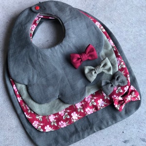 May include: Three layered baby bibs in shades of gray and burgundy. The top layer is a solid gray bib with scalloped edges. The middle layer is a gray bib with a burgundy floral print. The bottom layer is a burgundy bib with a white floral print. Each bib has a burgundy bow.