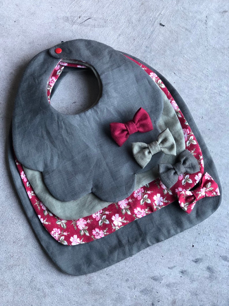 May include: Three layered baby bibs in shades of gray and burgundy. The top layer is a solid gray bib with scalloped edges. The middle layer is a gray bib with a burgundy floral print. The bottom layer is a burgundy bib with a white floral print. Each bib has a burgundy bow.
