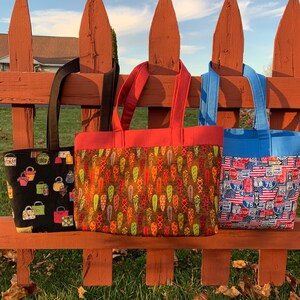 May include: Three tote bags hanging on a red wooden fence. The bag on the left has a black background with colourful handbags printed on it. The middle bag has a brown background with colourful leaves printed on it. The bag on the right has a blue background with red, white and blue highway signs printed on it.