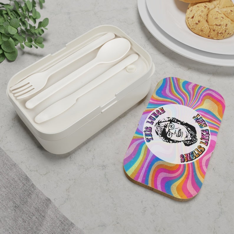 Puede incluir: Una fiambrera blanca con un dise&ntilde;o psicod&eacute;lico colorido en la tapa. La tapa presenta una cara de dibujos animados con el texto "This lunch smells like...