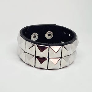 Op de afbeelding: Zwarte leren armband met twee rijen zilveren piramide studs. De armband heeft twee drukknoopsluitingen. Een opvallend punkrock-accessoire tegen een witte achtergrond.