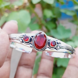 May include: A silver bangle bracelet with three red gemstone accents. The centre stone is oval-shaped and the two smaller stones are round.