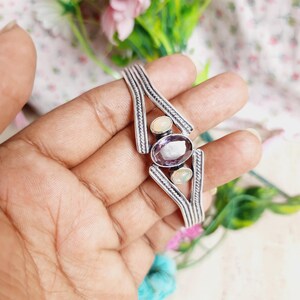 May include: Silver bangle bracelet with a geometric design featuring three oval-shaped gemstones: a purple amethyst and two white opals.