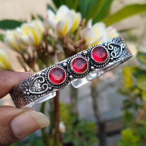 May include: Silver bangle bracelet with four red gemstone accents. The bracelet has an ornate design with floral and heart motifs.