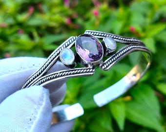 May include: Silver bangle bracelet featuring a purple amethyst gemstone and two moonstone gemstones. The bracelet has a braided design.