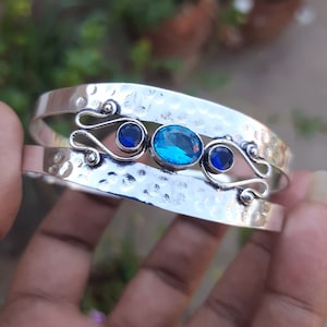 May include: Silver bangle bracelet with a hammered finish and three blue gemstones. The center stone is a larger oval shape, and the two smaller stones are round.