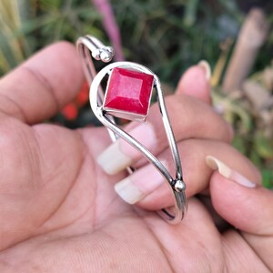 May include: A silver cuff bracelet with a striking, square, bright red gemstone. The bracelet has a minimalist design with a curved band and decorative details. The gemstone is the focal point, set in a simple bezel.