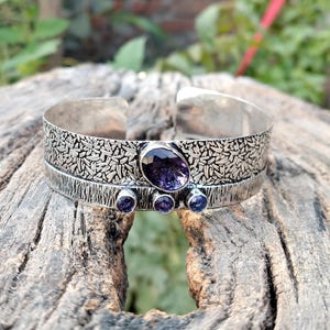 May include: A silver cuff bracelet with an oval purple gemstone and three smaller round purple gemstones. The bracelet has a textured leaf pattern on the top half and is displayed on a wooden surface.