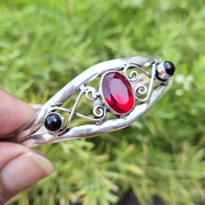 May include: A silver cuff bracelet featuring a central oval-cut red gemstone flanked by two smaller round black stones. The metal band displays intricate swirling wirework patterns that connect the stones within the open frame design.