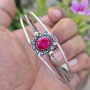 Puede incluir: Brazalete plateado con una gema roja rubí ovalada en el centro. La gema está engastada en un diseño floral. El brazalete tiene un diseño sencillo con un detalle de cuerda trenzada. Joyería.