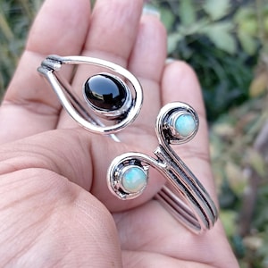 May include: A silver-toned ring and bracelet set. The ring features a black oval stone, while the bracelet has two light blue opal-like stones. The jewelry has a swirling, organic design.