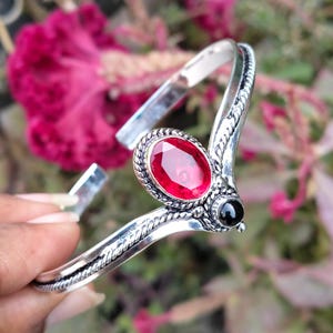 May include: Silver-toned cuff bracelet featuring a large oval red gemstone and a small black stone. The bracelet has a decorative rope-like design along the edges and is set against a blurred floral background.