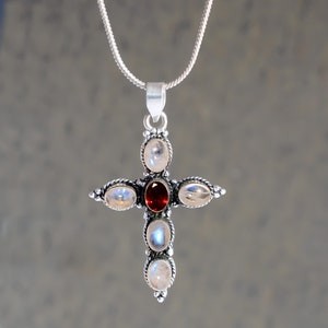 May include: A silver cross pendant featuring five oval-shaped gemstones. The central stone is red, and the four surrounding stones are white.