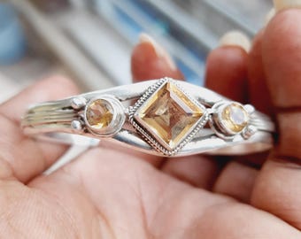 May include: A silver bangle bracelet with three yellow gemstones. The center stone is a square-shaped, faceted gemstone. The two smaller stones are round and are set on either side of the center stone.