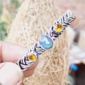 Op de afbeelding: Een zilveren armband met een gevlochten ontwerp en drie edelstenen. De middelste steen is een grote, iriserende blauwe steen, en de twee kleinere stenen zijn geel.