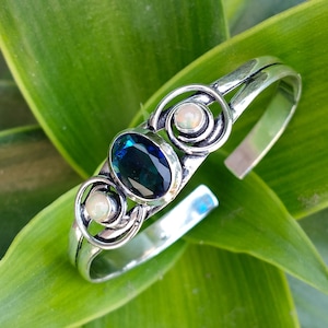 May include: Silver cuff bracelet featuring an oval, dark blue gemstone and two smaller, round, white gemstones. The bracelet has a decorative swirl design and is set against a green leaf background.