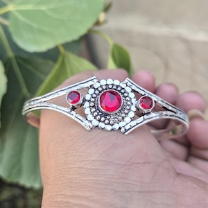 May include: A silver bracelet featuring three red gemstones. The central gemstone is encircled by a pattern of small silver beads. The bracelet has an open design with decorative edges.