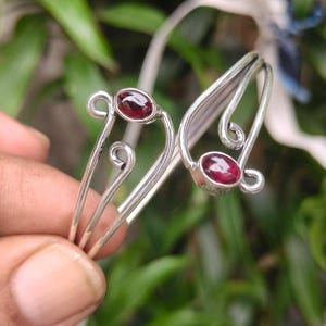 May include: A silver bracelet featuring two oval, deep red gemstones. The bracelet has a delicate, open design with swirling accents. The gemstones are set in a bezel setting, and the bracelet is shown against a blurred green background.