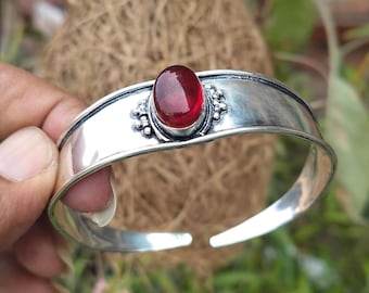 Könnte beinhalten: Ein silbernes Armreif-Armband mit einem roten Edelstein, der in einer silbernen Fassung sitzt. Das Armband hat ein schlichtes Design mit einem dekorativen Rand um den Edelstein.