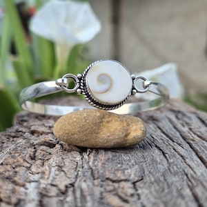 Op de afbeelding: Een zilveren armband met een centrale ronde Shiva-oog schelp inlay. De schelp is wit met een natuurlijk bruin spiraalpatroon. De zetting is omlijst door een gedraaide zilveren touwrand en verbonden met een gladde, gepolijste zilveren band.