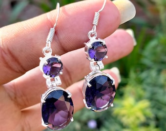 May include: A pair of silver drop earrings featuring two oval-cut, deep purple gemstones. The earrings have a simple design with a hook closure and a small, round gemstone above the larger oval stone. The gemstones are faceted, catching the light.