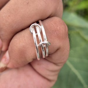 May include: A polished silver ring featuring a multi-strand design with three interconnected loops that overlap and wrap around the finger. The metal has a smooth, reflective surface and a continuous, fluid structure that creates a layered appearance.