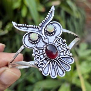 Puede incluir: Un brazalete de plata con diseño de búho, con una gema roja ovalada central y dos gemas verdes más pequeñas. El diseño del búho presenta detalles intrincados, sobre un fondo verde borroso.