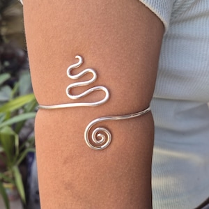 May include: A silver-colored arm cuff with a unique design. The cuff features a wavy, serpentine pattern on the upper arm and a spiral design on the lower arm. The jewelry is worn on the arm.