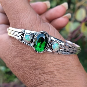 May include: A silver bangle bracelet featuring a green gemstone and two white opals. The bracelet has an ornate design.