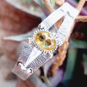 Op de afbeelding: Een zilveren armband met een getextureerde afwerking en een grote ovale gele edelsteen in het midden.