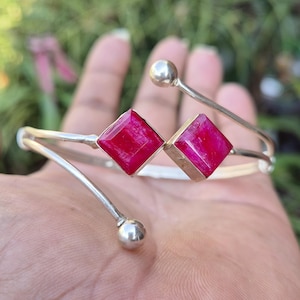 May include: A silver cuff bracelet with two square, vivid red gemstones. The bracelet has a minimalist design with a curved shape and small silver ball accents. The gemstones are set in a gold-toned metal. A piece of jewellery.