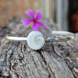 Könnte beinhalten: Ein silbernes Armreif-Armband mit einem weißen, kreisförmigen Mittelstück mit einem Spiralmuster. Das Armband liegt auf einer strukturierten Holzoberfläche, mit einer verschwommenen rosa Blume im Hintergrund. Das Design des Armbands ist schlicht und elegant.