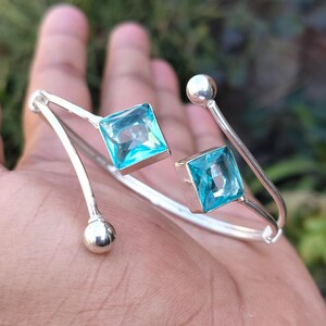 Pode incluir: Uma pulseira de punho prateada com duas pedras preciosas quadradas azuis claras. A pulseira tem um design minimalista com linhas curvas e pequenos detalhes esféricos. As pedras preciosas são definidas em molduras quadradas.