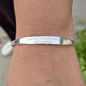 Puede incluir: Una pulsera de brazalete de metal plateado pulido con una inscripción grabada que dice (LOVER NEVER FAIL) seguida de la fecha (07/15/1983) en la superficie frontal de la banda, que tiene un diseño ligeramente curvado y cónico.