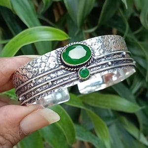 Pode incluir: Pulseira de punho de prata com textura martelada e duas pedras preciosas verdes. A pedra oval maior está centrada, com uma pedra redonda menor abaixo. A pulseira tem um design detalhado e texturizado.