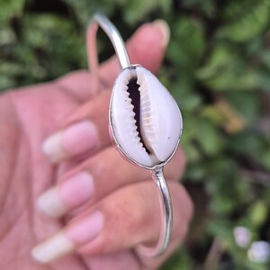 May include: A silver-toned metal bangle bracelet featuring a single, centered white cowrie shell. The shell has a smooth, rounded surface with a dark, serrated opening running down the middle, securely set within a polished metal frame.