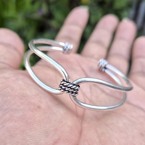 May include: A silver-toned cuff bracelet featuring two smooth, parallel bands that converge at the center. The center point is secured by a decorative, twisted rope-style metal knot. The ends of the cuff are finished with matching twisted rope details.