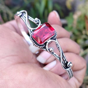 Puede incluir: Una pulsera plateada con una gran gema roja rectangular. La pulsera tiene un diseño calado adornado con patrones arremolinados y pequeños acentos redondos. La gema es el punto focal, engastada en un bisel detallado.