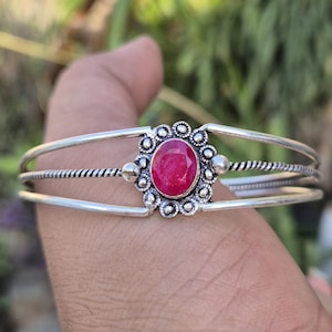 Könnte beinhalten: Silbernes Armreif-Armband mit einem leuchtenden, oval geschliffenen Rubin-Edelstein in der Mitte. Das Armband hat ein dreibändiges Design mit einem gedrehten Seildetail am mittleren Band. Der Rubin ist in eine dekorative, floral inspirierte Fassung gesetzt.
