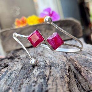 Può includere: Un bracciale a polsino argentato con due gemme quadrate rosso intenso. Il bracciale ha un design minimalista con una forma curva e piccole sfere argentate alle estremità. Le gemme sono incastonate in una montatura argentata.
