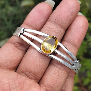 Op de afbeelding: Zilveren armband met een gele citrien edelsteen in het midden. De armband heeft drie banden en een stervormig ontwerp aan de uiteinden.