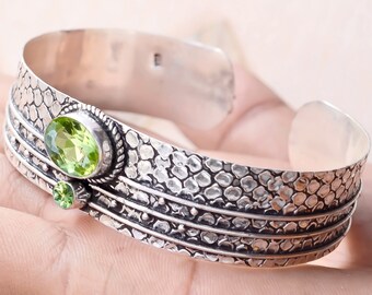 Könnte beinhalten: Silbernes Armreif-Armband mit strukturiertem Design und zwei grünen Peridot-Edelsteinen. Das Armband ist offen und verstellbar.