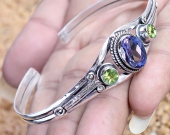 May include: A silver cuff bracelet featuring an oval, deep purple gemstone and two smaller, round, light green gemstones. The bracelet has an open design and is adorned with intricate metalwork details.