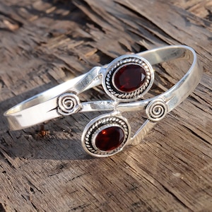 Op de afbeelding: Zilveren armband met twee rode edelsteen accenten. De armband heeft een delicaat, ingewikkeld ontwerp met draaiende details.