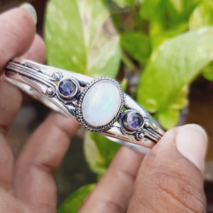 Può includere: Un braccialetto rigido in argento con una grande pietra di luna ovale bianca e due pietre di ametista ovali più piccole. Il braccialetto è decorato con intricati disegni in argento.
