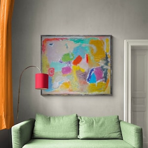 May include: Abstract painting with a colorful, textured design in shades of blue, green, yellow, pink, and orange. The painting is framed in a simple wooden frame and hangs on a light gray wall above a green sofa.