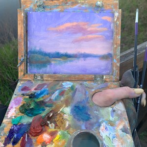 May include: A wooden artist's palette with a variety of colourful oil paints, a small metal container of water, and a thumbprint in the wet paint. The palette is open and a small painting of a purple and pink sunset over a lake is visible in the background.