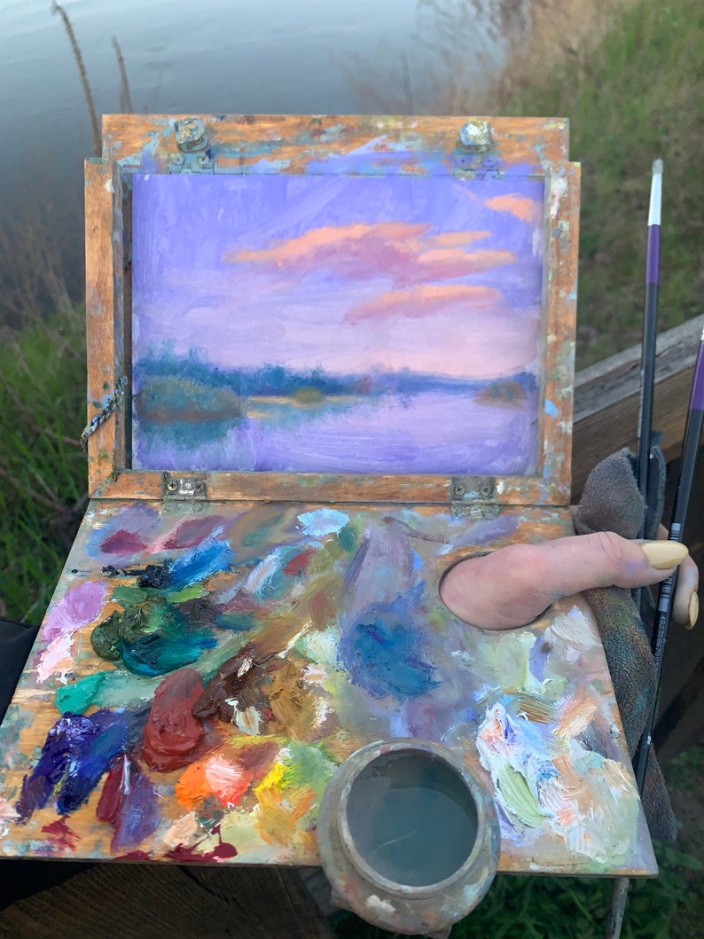 May include: A wooden artist's palette with a variety of colourful oil paints, a small metal container of water, and a thumbprint in the wet paint. The palette is open and a small painting of a purple and pink sunset over a lake is visible in the background.