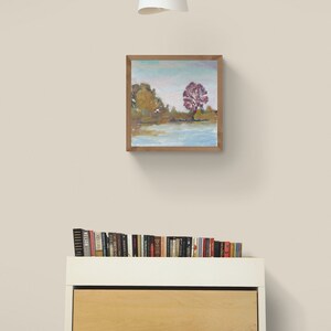 Puede incluir: Una pintura enmarcada de un paisaje con un solo &aacute;rbol rojo en el centro, rodeado de &aacute;rboles verdes y un lago azul. La pintura est&aacute; en un marco de madera y est&aacute; colgada en una pared blanca.